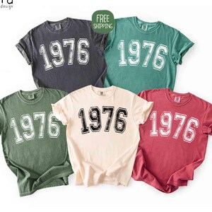 May include: Five short-sleeved t-shirts in various colors: charcoal, teal, olive green, cream, and maroon. Each shirt features the number "1976" in a distressed, collegiate-style font. The shirts are from "eyra design".