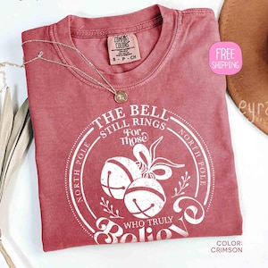 Op de afbeelding: Karmizijnrood T-shirt met een ronde afbeelding met de tekst "The Bell Still Rings For Those Who Truly Believe". Het ontwerp bevat bellen en "North Pole". Een gouden ketting ligt op het shirt.