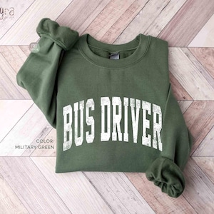 Può includere: Una felpa girocollo verde militare con la scritta BUS DRIVER stampata in un carattere bianco dall'aspetto usurato sul petto. Il capo presenta maniche lunghe con polsini a coste e uno scollo a coste, con un design semplice e funzionale.