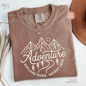 May include: Espresso-colored t-shirt with a white mountain graphic and the word "Adventure". The shirt also features the text "Explore More, Worry Less". A silver necklace and a brown hat are also visible.