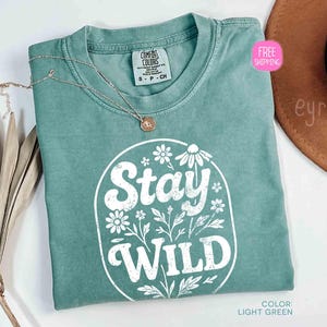 May include: Light green t-shirt with the text "Stay Wild" in a white floral design. The shirt is a Comfort Colors brand, size S-P-CH. A silver necklace with a pendant is on the shirt. The color is light green.