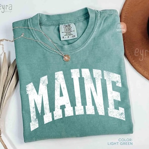 May include: A light green t-shirt with the word "MAINE" in large, distressed white letters. A gold necklace with a pendant rests on the shirt. The shirt is labeled "Comfort Colors" and is a size small.