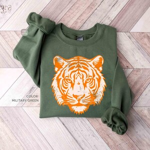 Può includere: Una felpa girocollo verde militare con una grande grafica arancione e bianca dall'aspetto vissuto raffigurante un muso di tigre al centro. Il capo presenta maniche lunghe con polsini a coste e scollo a coste, con il testo COLOR: MILITARY GREEN.