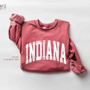 Könnte beinhalten: Ein scharlachrotes Langarm-Sweatshirt mit Rundhalsausschnitt und dem Wort INDIANA in einer großen, weißen, abgenutzten Schriftart im College-Stil auf der Brust. Das Kleidungsstück hat einen gerippten Ausschnitt und Bündchen, mit dem Text eyra design.