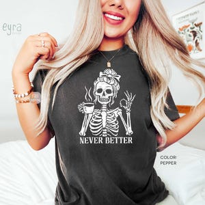 May include: A pepper-coloured t-shirt featuring a white graphic of a skeleton with a messy bun holding a coffee cup and making an okay hand gesture. The text below the skeleton reads NEVER BETTER in a bold, white, capitalised serif font.