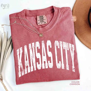 May include: Crimson-colored t-shirt with the text "KANSAS CITY" in distressed white lettering. A gold necklace with a pendant rests on the shirt. The shirt is a Comfort Colors brand, size S-P-CH.