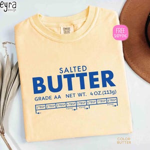 May include: A light yellow t-shirt with the word "BUTTER" in large blue letters. The text "SALTED" is above the word "BUTTER". Below the word "BUTTER" is the text "GRADE AA NET WT. 4 OZ.(113g)". There is a measurement chart below the text.