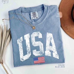 Puede incluir: Una camiseta azul vaquero con la palabra "USA" en letras blancas grandes y desgastadas. Debajo de las letras hay una peque&ntilde;a bandera americana. Un collar dorado descansa sobre la camisa. La camiseta es de la marca Comfort Colors.