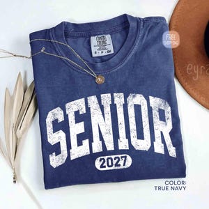 May include: A true navy blue t-shirt with the word "SENIOR" in distressed white lettering and the year "2027" below. A gold necklace rests on the shirt. The shirt is from Comfort Colors.