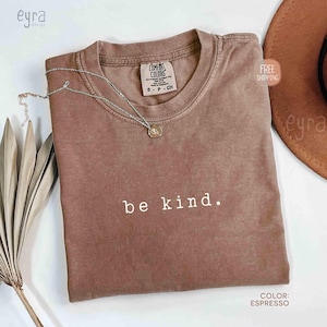 May include: Espresso-colored t-shirt with the words "be kind" in white. The shirt has a crew neck and a Comfort Colors tag. A silver necklace with a pendant is draped on the shirt. A brown hat is visible in the background.