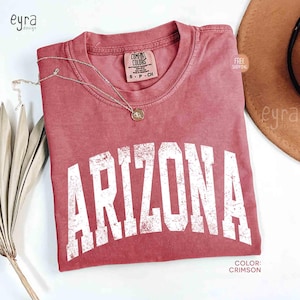 May include: A crimson short-sleeve t-shirt with a crew neckline featuring the word ARIZONA printed in large, distressed white block letters across the chest. The shirt has a Comfort Colors brand label at the inner neck and a small gold-toned necklace.
