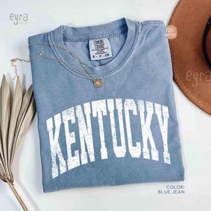 Könnte beinhalten: Ein blaues Jeans-T-Shirt mit dem Wort "KENTUCKY" in Weiß auf der Vorderseite aufgedruckt. Eine goldene Halskette mit Anhänger liegt auf dem Shirt. Das Shirt ist von der Marke Comfort Colors, Größe S-CH.
