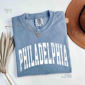 May include: A blue jean coloured short-sleeve t-shirt with a crew neckline and a distressed white arched graphic that reads PHILADELPHIA. The shirt features a Comfort Colors brand label at the inner neck and is paired with a delicate gold chain necklace.