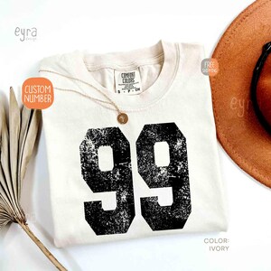 K&ouml;nnte beinhalten: Elfenbeinfarbenes T-Shirt mit der Nummer 99 in verblichenem schwarzem Druck. Eine goldene Halskette mit Anh&auml;nger liegt auf dem Shirt. Das Shirt ist mit "Comfort Colors" gekennzeichnet. Der Text "Custom Number" befindet sich auf einem orangefarbenen Aufkleber.