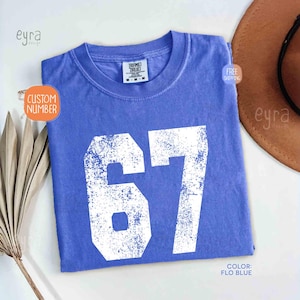 K&ouml;nnte beinhalten: Ein blaues T-Shirt mit der Nummer 67 in wei&szlig;er, abgenutzter Schrift. Das Shirt hat einen Rundhalsausschnitt und kurze &Auml;rmel. Die Farbe wird als "Flo Blue" bezeichnet. Das Shirt ist gefaltet und auf einer wei&szlig;en Oberfl&auml;che ausgestellt.