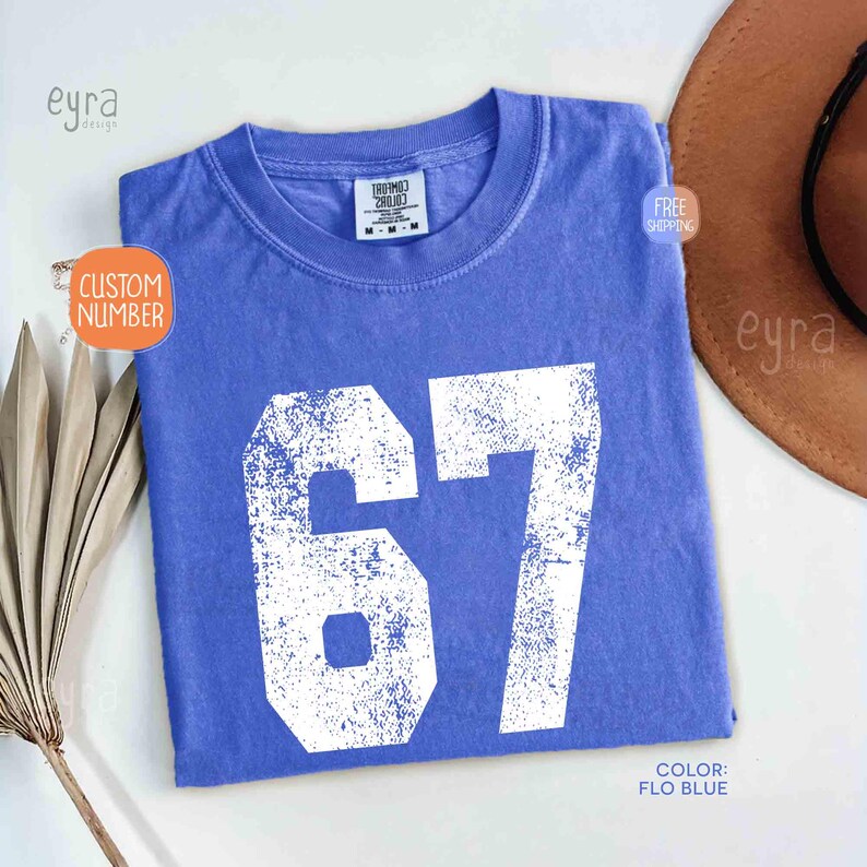 K&ouml;nnte beinhalten: Ein blaues T-Shirt mit der Nummer 67 in wei&szlig;er, abgenutzter Schrift. Das Shirt hat einen Rundhalsausschnitt und kurze &Auml;rmel. Die Farbe wird als "Flo Blue" bezeichnet. Das Shirt ist gefaltet und auf einer wei&szlig;en Oberfl&auml;che ausgestellt.