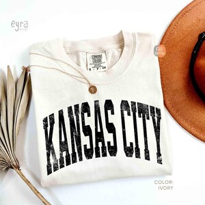 May include: Ivory-colored t-shirt with the text "KANSAS CITY" in distressed black lettering. A gold necklace with a pendant rests on the shirt. A brown hat and dried plant are also in the image. The shirt is labeled "Comfort Colors".