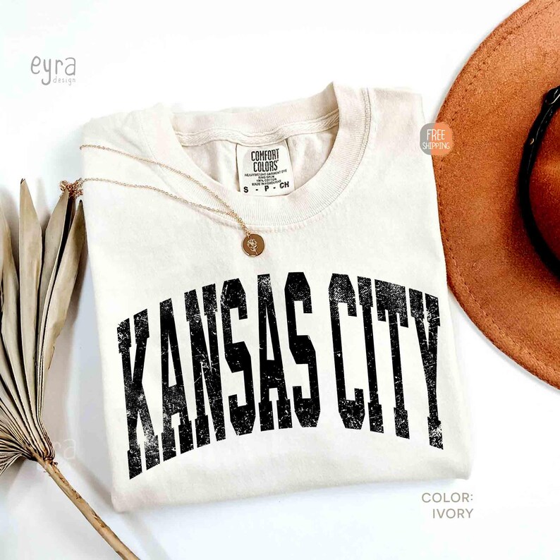 May include: Ivory-colored t-shirt with the text "KANSAS CITY" in distressed black lettering. A gold necklace with a pendant rests on the shirt. A brown hat and dried plant are also in the image. The shirt is labeled "Comfort Colors".