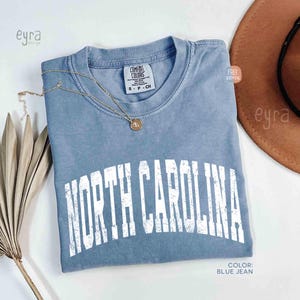 K&ouml;nnte beinhalten: Ein blaues T-Shirt in Jeansoptik mit Rundhalsausschnitt und kurzen &Auml;rmeln. Auf der Brust befindet sich der Schriftzug NORTH CAROLINA in einer abgenutzten wei&szlig;en Schriftart. Eine goldfarbene Halskette mit kleinem Anh&auml;nger liegt am Kragen.