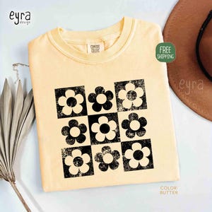 May include: Butter-colored t-shirt with a black and white floral design. The design features nine flowers in a grid pattern. The shirt is a Comfort Colors brand.