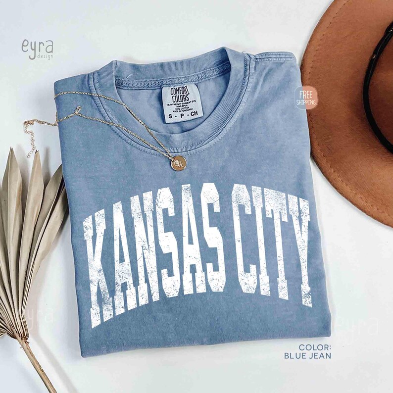 May include: A blue jean colored t-shirt with the text "KANSAS CITY" in white, distressed lettering. A gold necklace with a pendant rests on the shirt. The shirt is folded, and a brown hat is visible in the background.