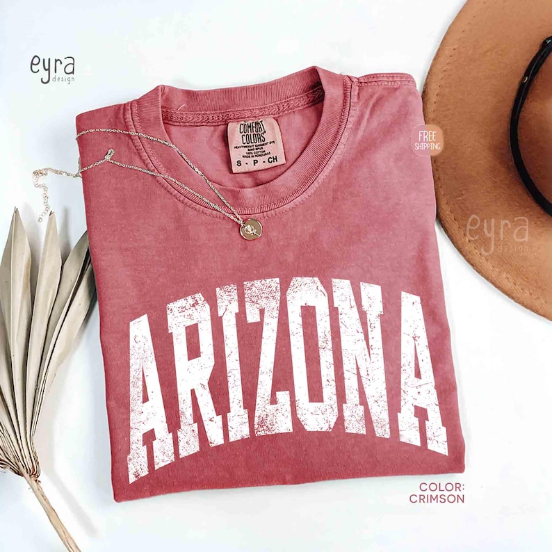 May include: A crimson short-sleeve t-shirt with a crew neckline featuring the word ARIZONA printed in large, distressed white block letters across the chest. The shirt has a Comfort Colors brand label at the inner neck and a small gold-toned necklace.