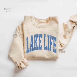 以下が含まれることがあります： 胸元に掠れた青いフォントでLAKE LIFEの文字がプリントされた、サンドカラーのクルーネックトレーナー。袖口と襟元はリブ編みになっており、長袖でシンプルかつ機能的なデザインの衣類です。