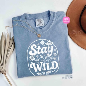 May include: A blue jean colored t-shirt with the words "Stay Wild" in white, surrounded by floral designs. The shirt has a crew neck and short sleeves. A gold necklace with a pendant is draped on the shirt.