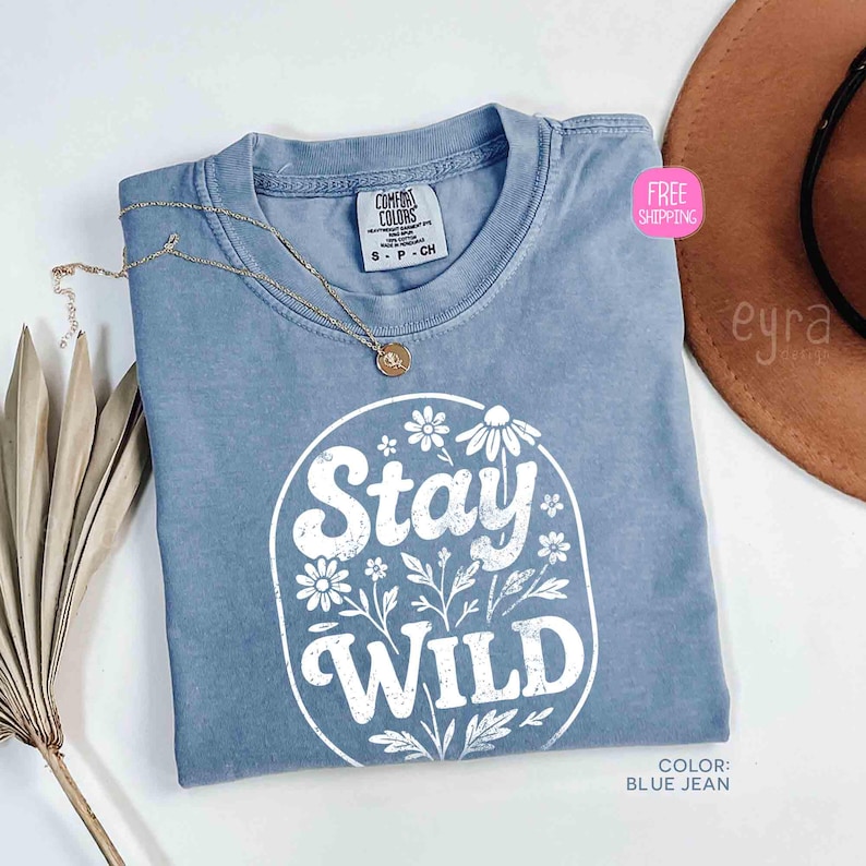 May include: A blue jean colored t-shirt with the words "Stay Wild" in white, surrounded by floral designs. The shirt has a crew neck and short sleeves. A gold necklace with a pendant is draped on the shirt.