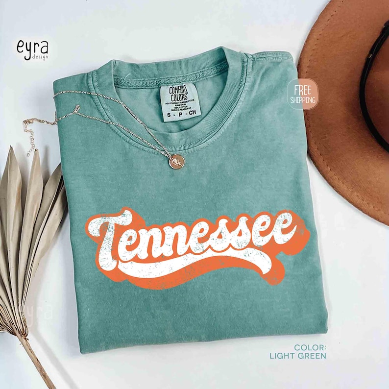 May include: A light green t-shirt with the word "Tennessee" in white retro font over an orange curved banner. The shirt has a crew neck and short sleeves. A silver necklace with a pendant is on the shirt.