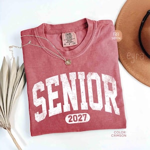 May include: Crimson-colored t-shirt with the word "SENIOR" in large white block letters and the year "2027" underneath. A gold necklace with a pendant rests on the shirt. The shirt is a Comfort Colors brand.