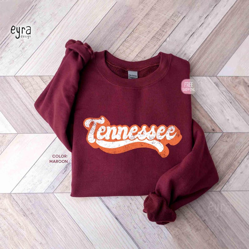 K&ouml;nnte beinhalten: Ein kastanienbraunes Sweatshirt mit Rundhalsausschnitt und dem Wort Tennessee in einer wei&szlig;en, abgenutzten Retro-Schriftart auf der Brust. Das Kleidungsst&uuml;ck hat lange &Auml;rmel, einen gerippten Kragen und ein sichtbares Gildan-Markenetikett am Halsausschnitt.