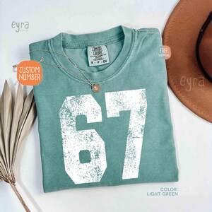 K&ouml;nnte beinhalten: Hellgr&uuml;nes T-Shirt mit der wei&szlig;en, verwitterten Zahl 67. Das Shirt hat einen Rundhalsausschnitt und kurze &Auml;rmel. Eine silberne Halskette mit Anh&auml;nger liegt auf dem Shirt. Der Text "CUSTOM NUMBER" befindet sich auf einem Aufkleber.