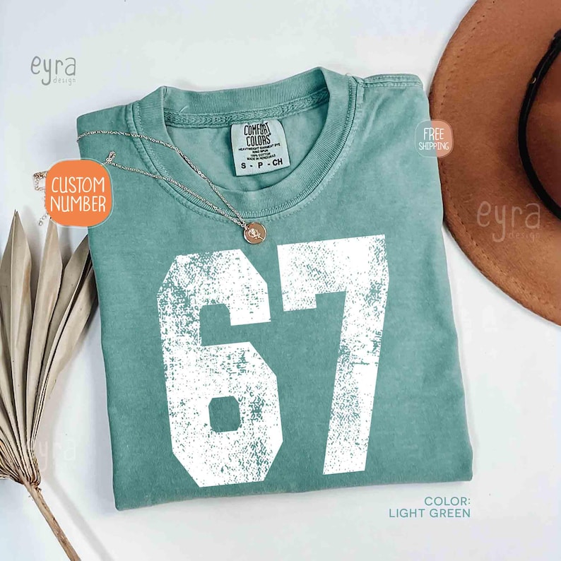 K&ouml;nnte beinhalten: Hellgr&uuml;nes T-Shirt mit der wei&szlig;en, verwitterten Zahl 67. Das Shirt hat einen Rundhalsausschnitt und kurze &Auml;rmel. Eine silberne Halskette mit Anh&auml;nger liegt auf dem Shirt. Der Text "CUSTOM NUMBER" befindet sich auf einem Aufkleber.