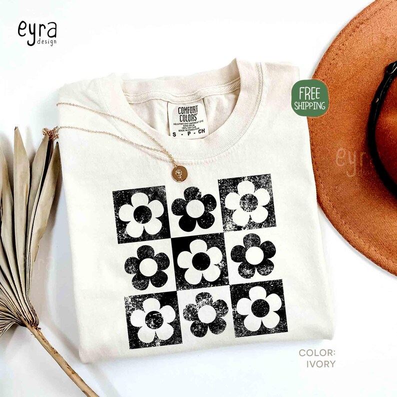 May include: Ivory-colored t-shirt featuring a black and white floral pattern. The shirt has a crew neck and short sleeves. A gold necklace with a pendant rests on the shirt. The shirt is labeled "Comfort Colors".