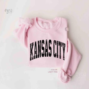May include: Pale pink sweatshirt featuring the words "KANSAS CITY" in distressed black font. The crew neck top has long sleeves. The colour is light pink.