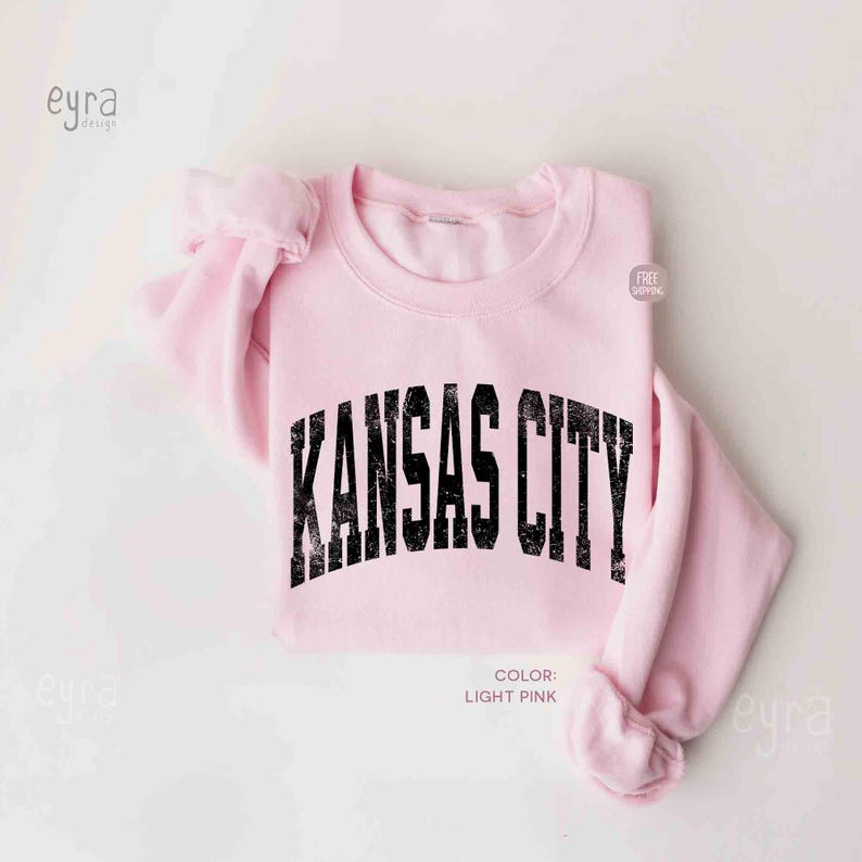 May include: Pale pink sweatshirt featuring the words "KANSAS CITY" in distressed black font. The crew neck top has long sleeves. The colour is light pink.