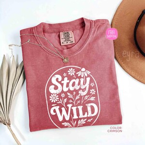 May include: Crimson-colored t-shirt with the words "Stay Wild" in white, surrounded by floral designs. A gold necklace rests on the shirt. The shirt is a Comfort Colors brand, size S-P-CH.