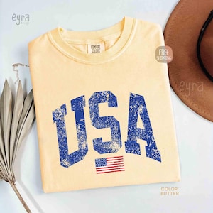 Puede incluir: Camiseta de color mantequilla con la palabra "USA" en letras azules desgastadas y una peque&ntilde;a bandera americana. La camiseta tiene cuello redondo y mangas cortas. La camiseta es de la marca Comfort Colors.