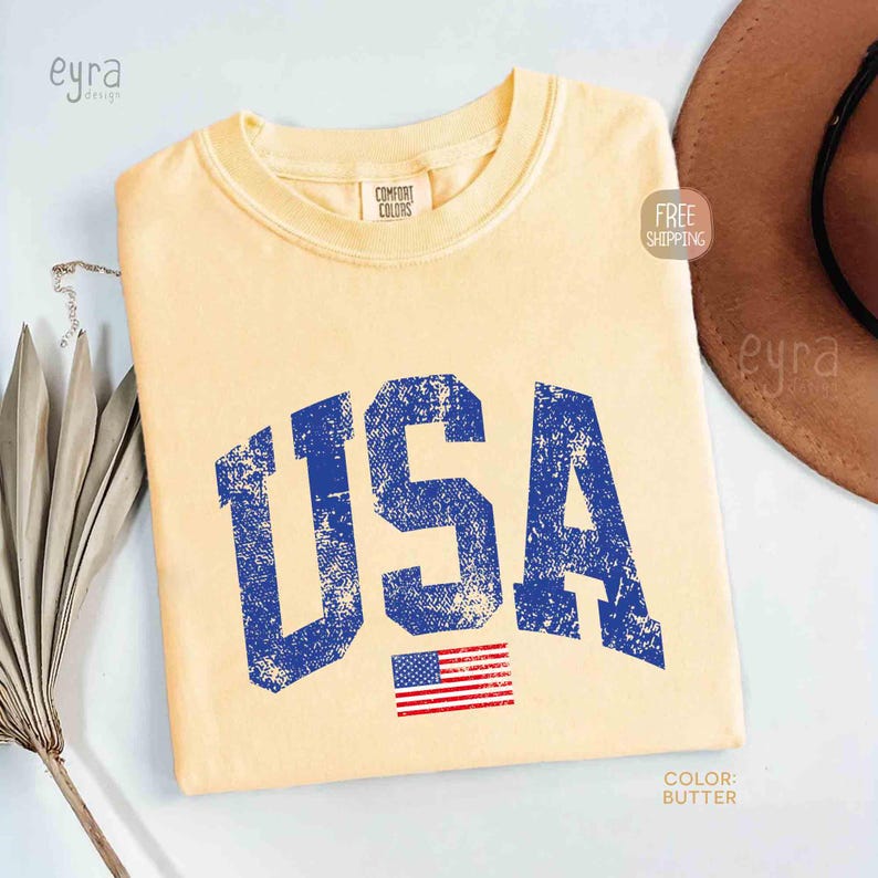 Puede incluir: Camiseta de color mantequilla con la palabra "USA" en letras azules desgastadas y una peque&ntilde;a bandera americana. La camiseta tiene cuello redondo y mangas cortas. La camiseta es de la marca Comfort Colors.