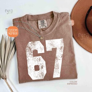 K&ouml;nnte beinhalten: Espressofarbenes T-Shirt mit der Nummer 67 in wei&szlig;er, verblichener Schrift. Das Shirt hat einen Rundhalsausschnitt und kurze &Auml;rmel. Eine silberne Halskette mit Anh&auml;nger liegt auf dem Shirt. Das Etikett lautet "Comfort Colors".