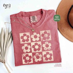May include: Crimson-colored t-shirt with a retro floral pattern. The shirt features a grid of nine flowers in a lighter shade, with the text "COMFORT COLORS" on the tag. A silver necklace with a pendant is draped on the shirt.