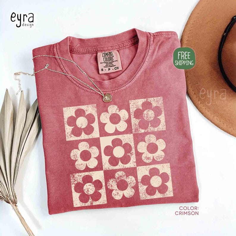 May include: Crimson-colored t-shirt with a retro floral pattern. The shirt features a grid of nine flowers in a lighter shade, with the text "COMFORT COLORS" on the tag. A silver necklace with a pendant is draped on the shirt.