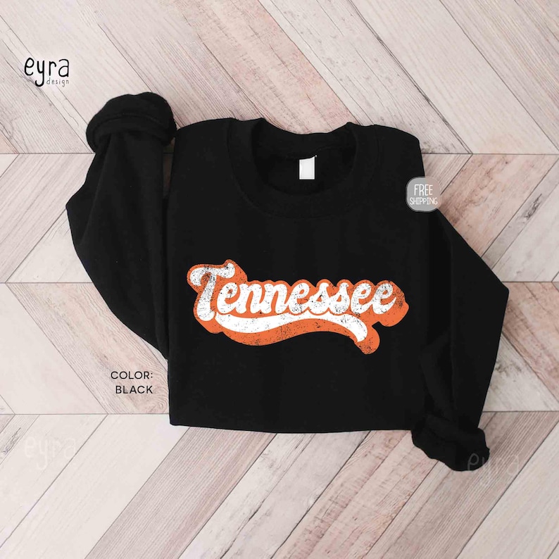 K&ouml;nnte beinhalten: Ein schwarzes Langarm-Sweatshirt mit Rundhalsausschnitt, das in der Mitte der Brust das Wort Tennessee in einer wei&szlig;en, abgenutzten Schreibschrift zeigt. Der Text ist mit einer dicken, orangefarbenen, texturierten Grafik umrandet.