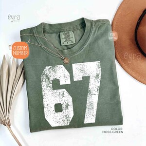 K&ouml;nnte beinhalten: Moosgr&uuml;nes T-Shirt mit der Nummer 67 in wei&szlig;er, verblichener Schrift. Das Shirt ist von der Marke Comfort Colors, Gr&ouml;&szlig;e S-P-CH. Eine goldene Halskette mit Anh&auml;nger liegt auf dem Shirt. Das Wort "Custom Number" steht auf einem Etikett.