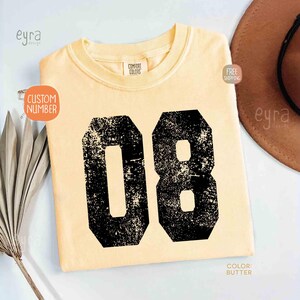 K&ouml;nnte beinhalten: Ein hellgelbes T-Shirt mit der Nummer "08" in verblichenem Schwarz aufgedruckt. Das T-Shirt hat einen Rundhalsausschnitt und kurze &Auml;rmel. Der Text "CUSTOM NUMBER" befindet sich auf einem Aufkleber auf dem Shirt. Die Farbe des Shirts ist "BUTTER".