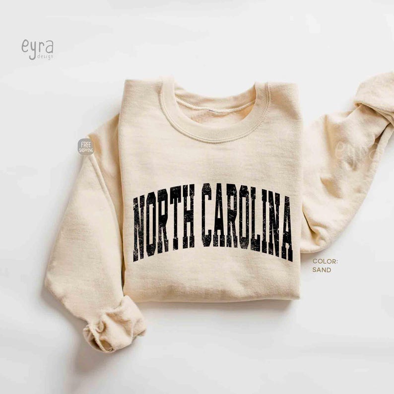 Może przedstawiać: Bluza z okrągłym dekoltem w kolorze piaskowym z napisem NORTH CAROLINA nadrukowanym czarną, postarzaną, łukowatą czcionką na klatce piersiowej. Odzież posiada długie rękawy oraz ściągacze przy kołnierzu, mankietach i u dołu.