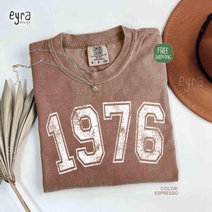 May include: Espresso-colored t-shirt with the year "1976" printed in white block letters. The shirt has a crew neck and a small tag on the collar. A silver necklace with a pendant is draped on the shirt. A brown hat is visible in the background.