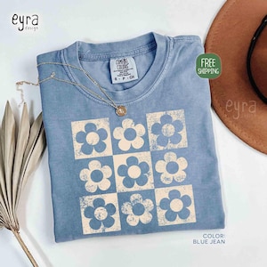 May include: Blue jean-colored t-shirt with a floral pattern. The shirt features a grid of nine flowers in a light beige color. The shirt has a crew neck and short sleeves. The tag reads "Comfort Colors". A gold necklace is draped over the shirt.