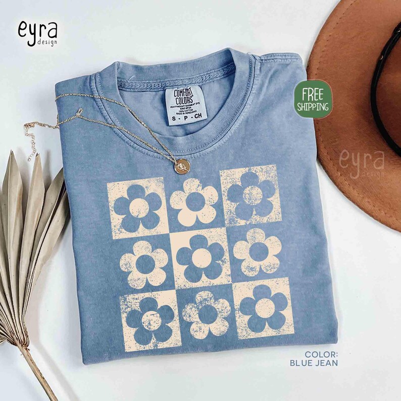 May include: Blue jean-colored t-shirt with a floral pattern. The shirt features a grid of nine flowers in a light beige color. The shirt has a crew neck and short sleeves. The tag reads "Comfort Colors". A gold necklace is draped over the shirt.